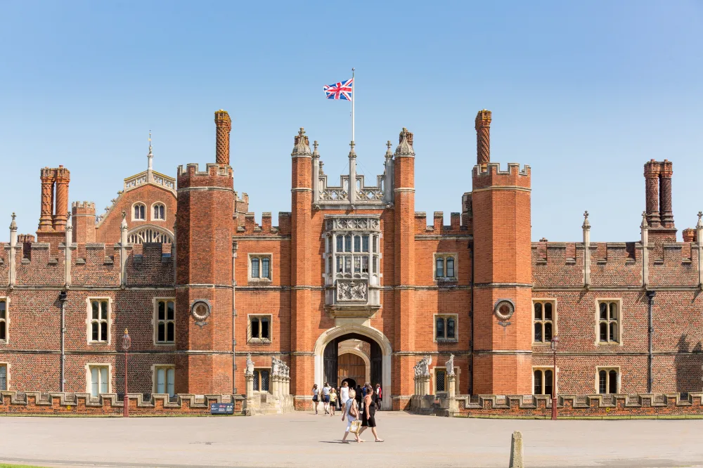 Image looks Hampton Court Palace face on underneath a clear sky. A United Kingdom flag flies high above the castle, and the facade of the castle in orange brick shines bright.