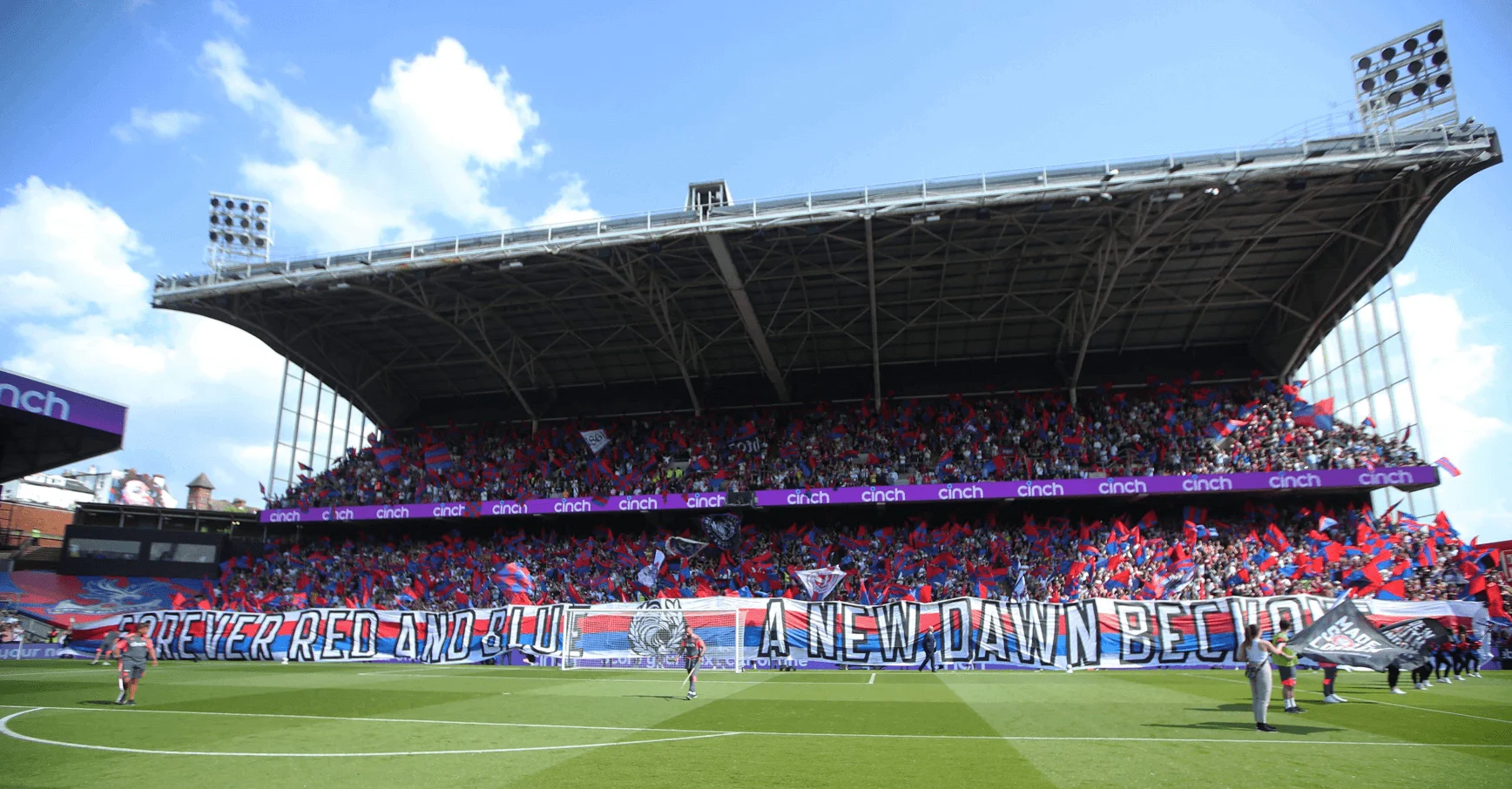A football stadium stand is packed with fans flying flags high behind the goal. A blue sky shines overhead. A banner lines the lower stand and reads 'Forever red and blue, a new dawn beckons'.