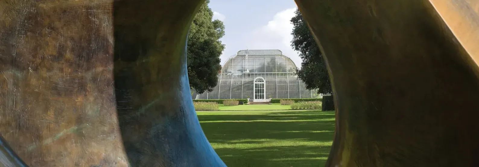 A Victorian glasshouse at Kew Gardens viewed through the curved forms of a bronze sculpture by Henry Moore, framed by green lawns and trees.