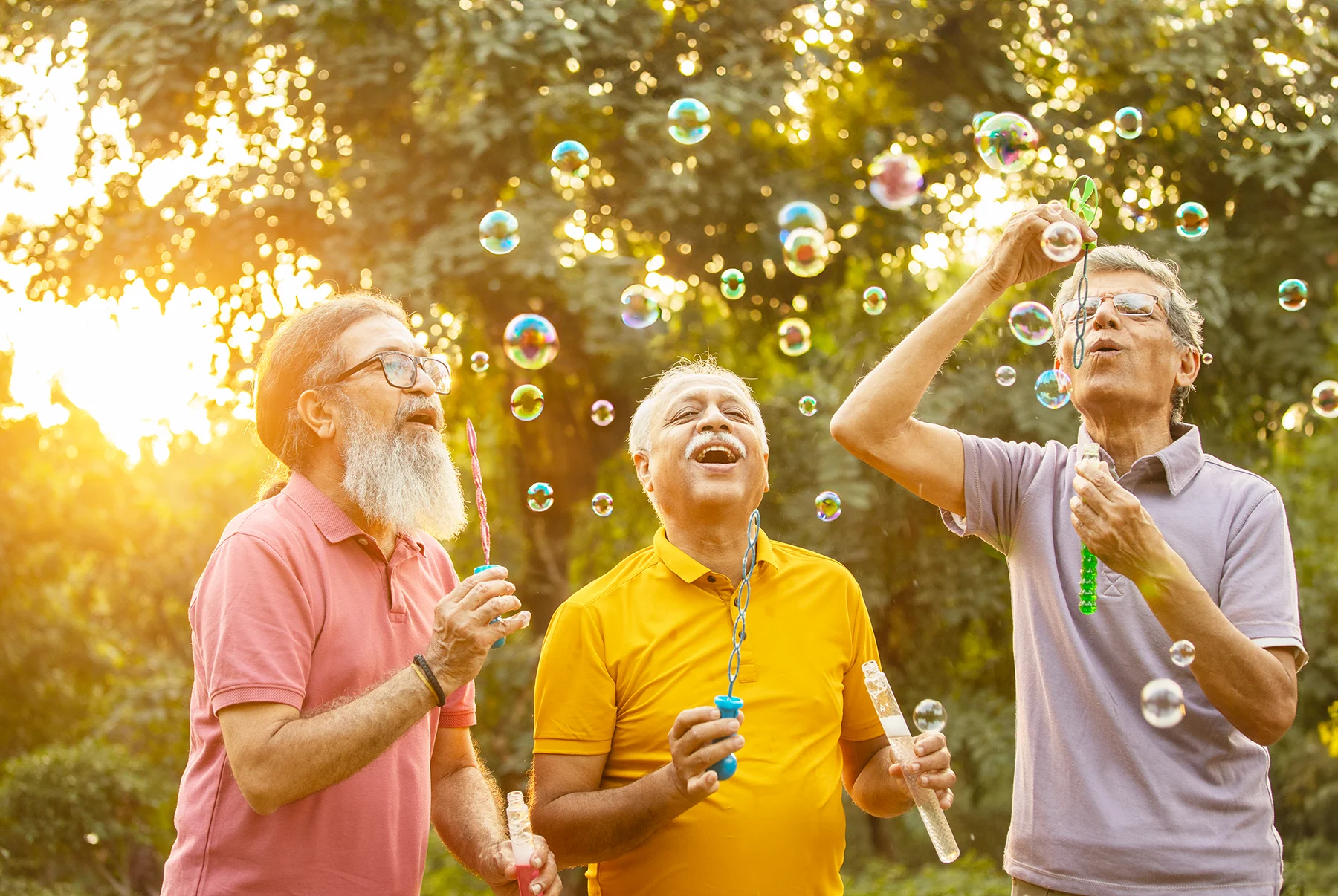 3 mature South Asian men laughing and blowing bubbles outside on a sunny day.