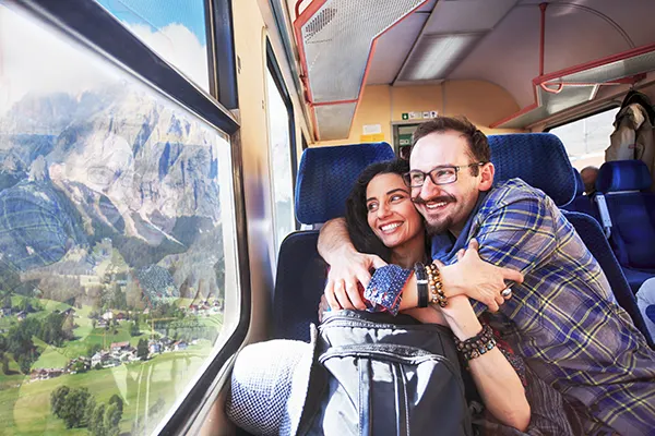 A man and woman smiling and hugging on a train, looking out the window at mountains and green fields. 