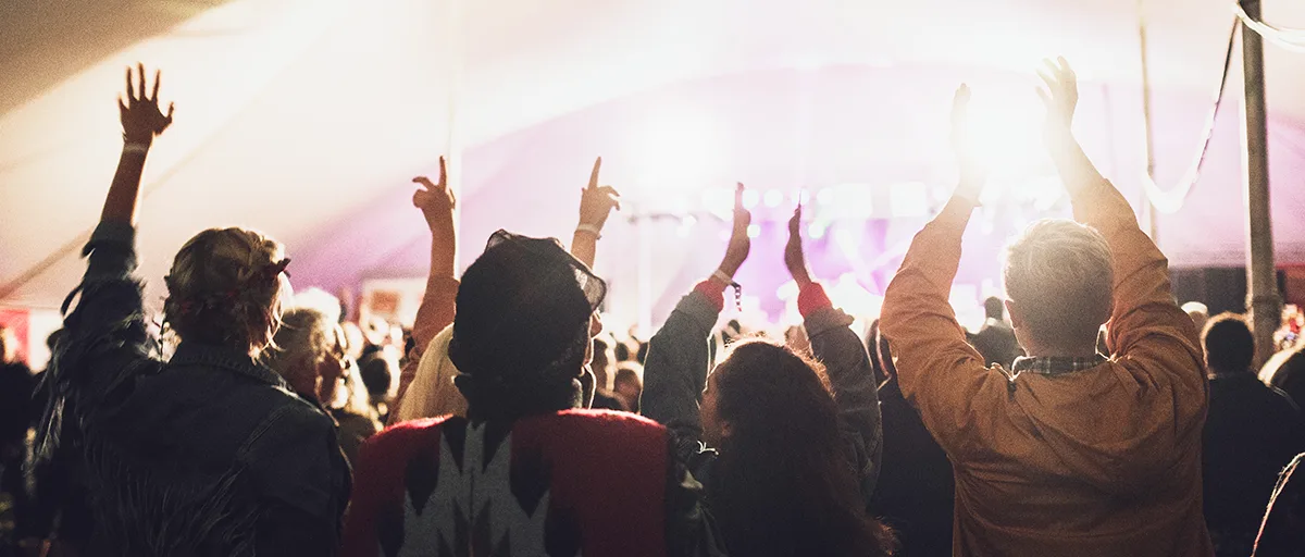A crowd of festival-goers with hands raised inside a large tent, bathed in warm and purple stage lighting.