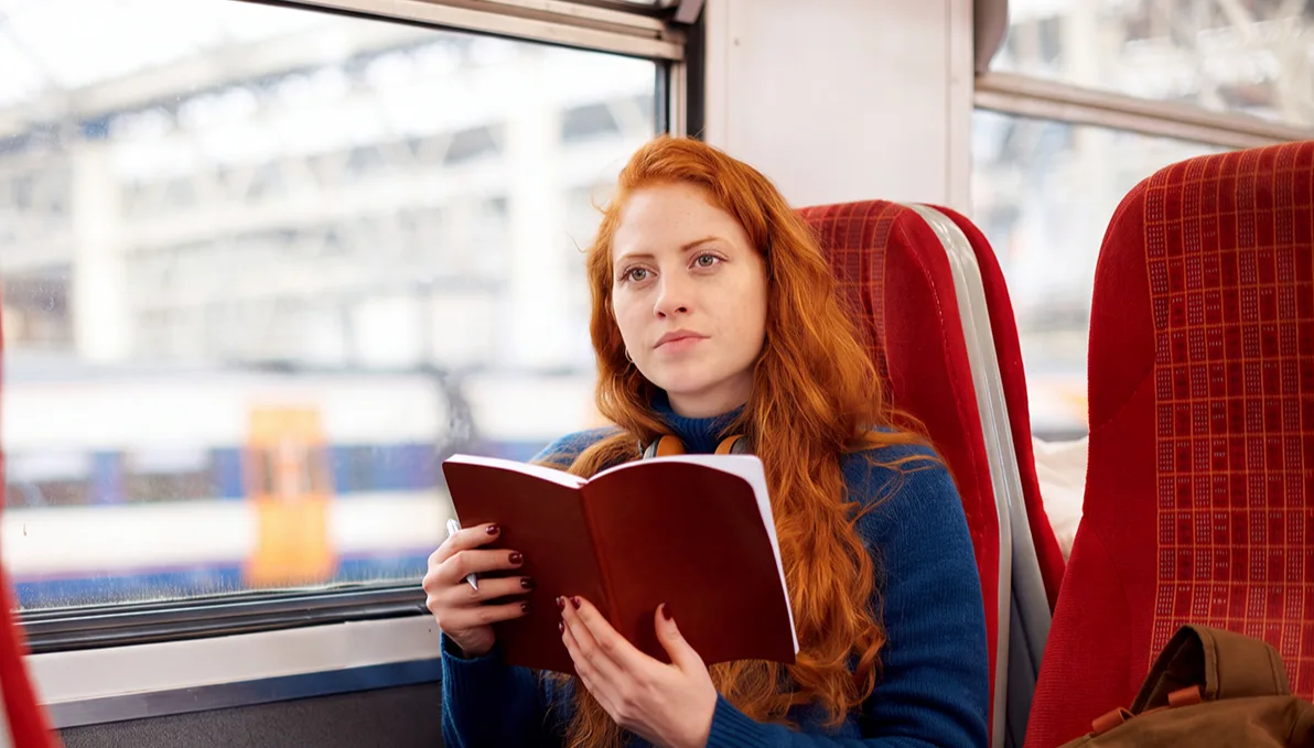 A young red-haired woman sits on a train holding an open notebook, wearing a blue jumper, with a rucksack beside her.