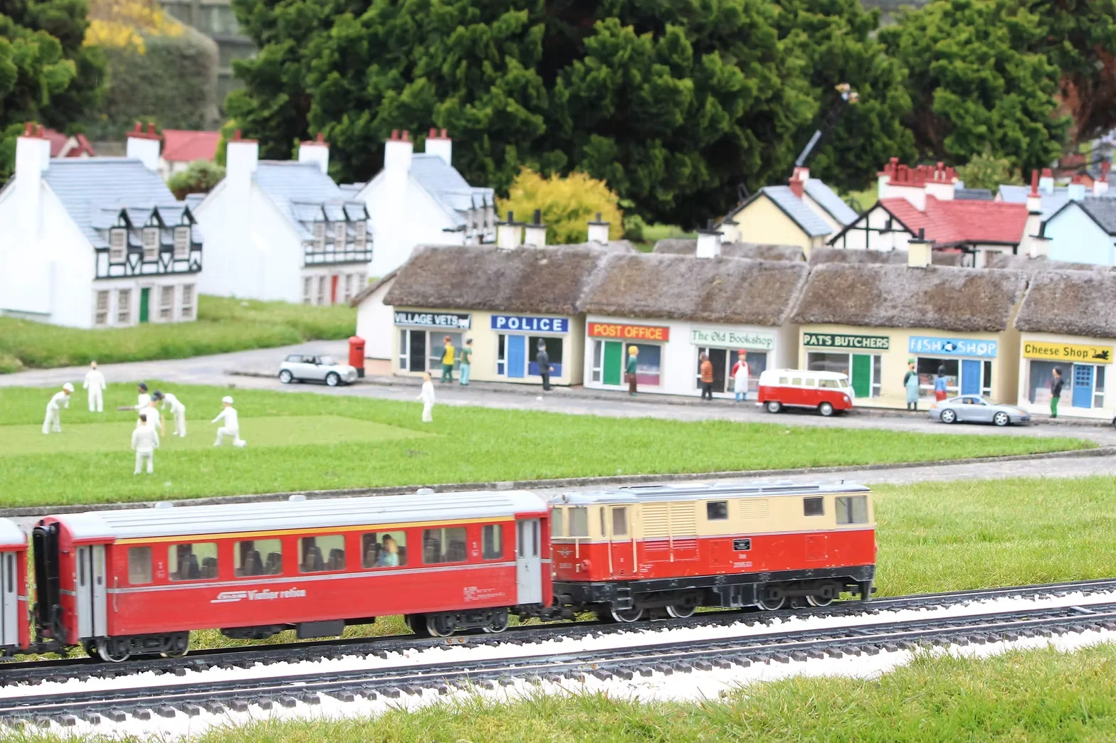A model village, with a model train crossing a village green with toy cricketers playing, and a parade of shops behind.