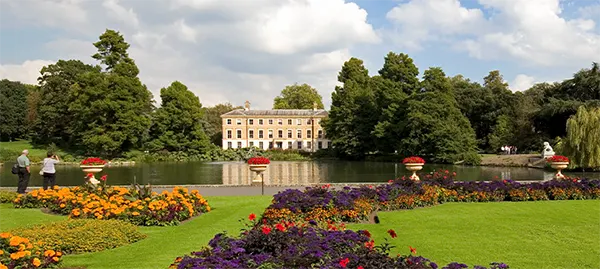 Historic mansion at Kew Gardens reflected in a lake, surrounded by manicured lawns with colourful flower beds under a cloudy sky.