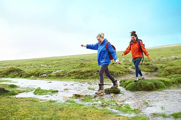Two hikers in blue and orange jackets crossing a stream on stepping stones in a green moorland landscape.