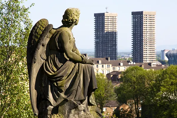 A statue of an angel on a hill, with two large tower blocks in the distance. 