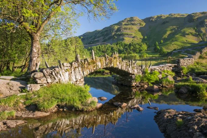 An old stone bridge connects two areas of green foliage over a quaint and still stream. Blue skies and green mountains in the background.