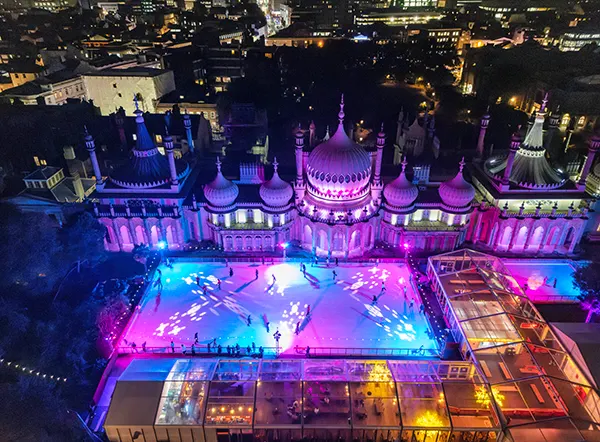 Aerial, nighttime view of an outdoor ice rink and a royal palace with turrets and minarets, bathed in purple and blue light.