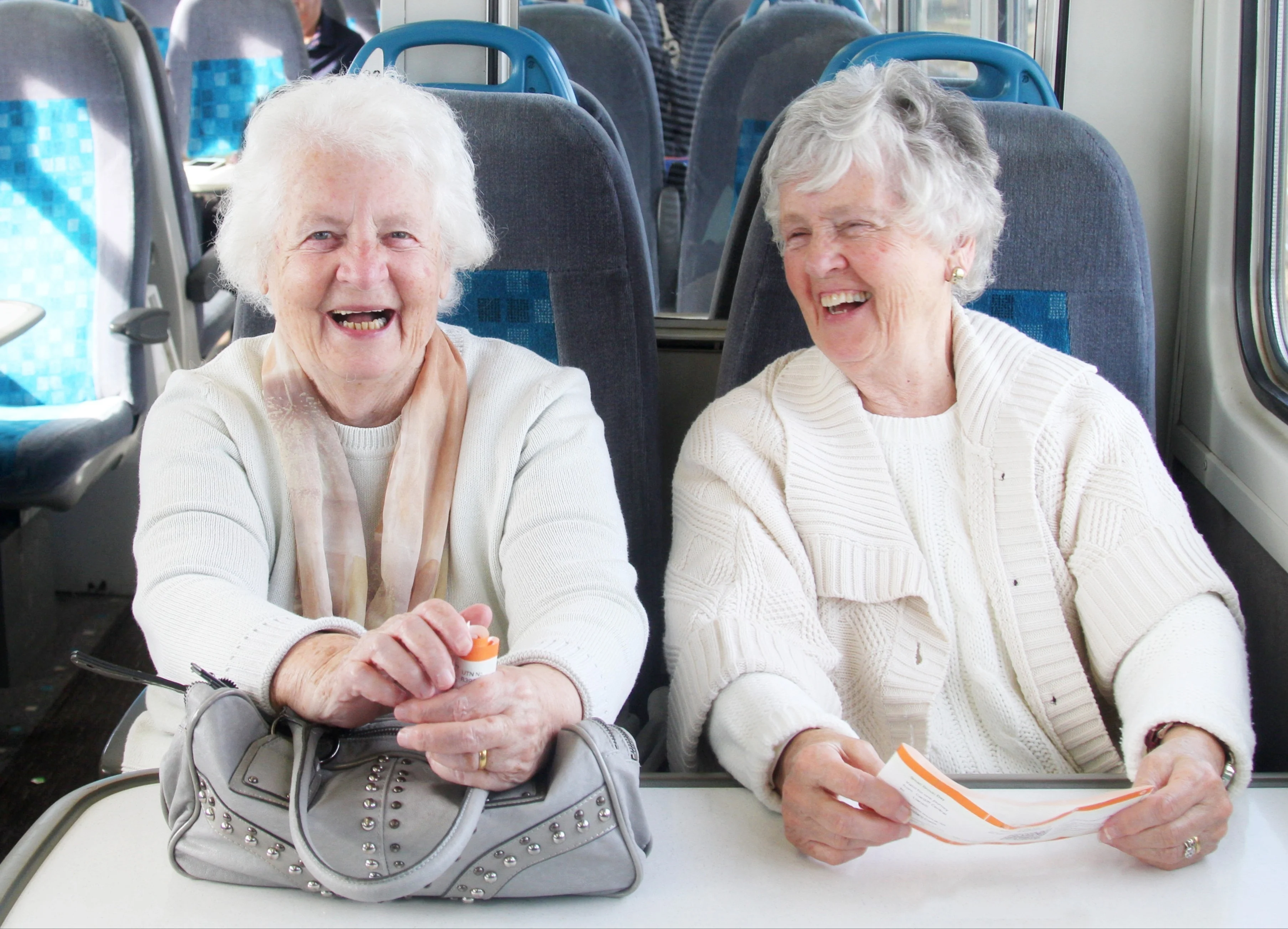 2 elderly white women with white hair are sitting next to each other at a train table, holding train tickets in their hands and laughing. 