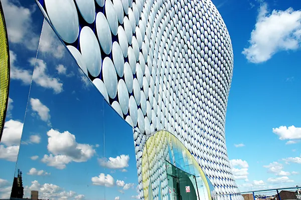 A curved building covered in white circular panels, with blue sky and clouds reflected in a mirrored surface on its side.