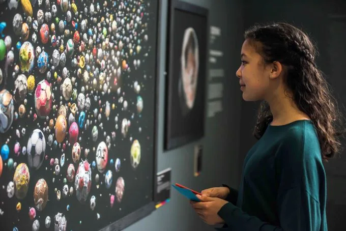 Young girl with curly dark brown hair and plait wearing sea-green long sleeve top gazes at artwork comprised of multicoloured footballs set on a black backdrop.