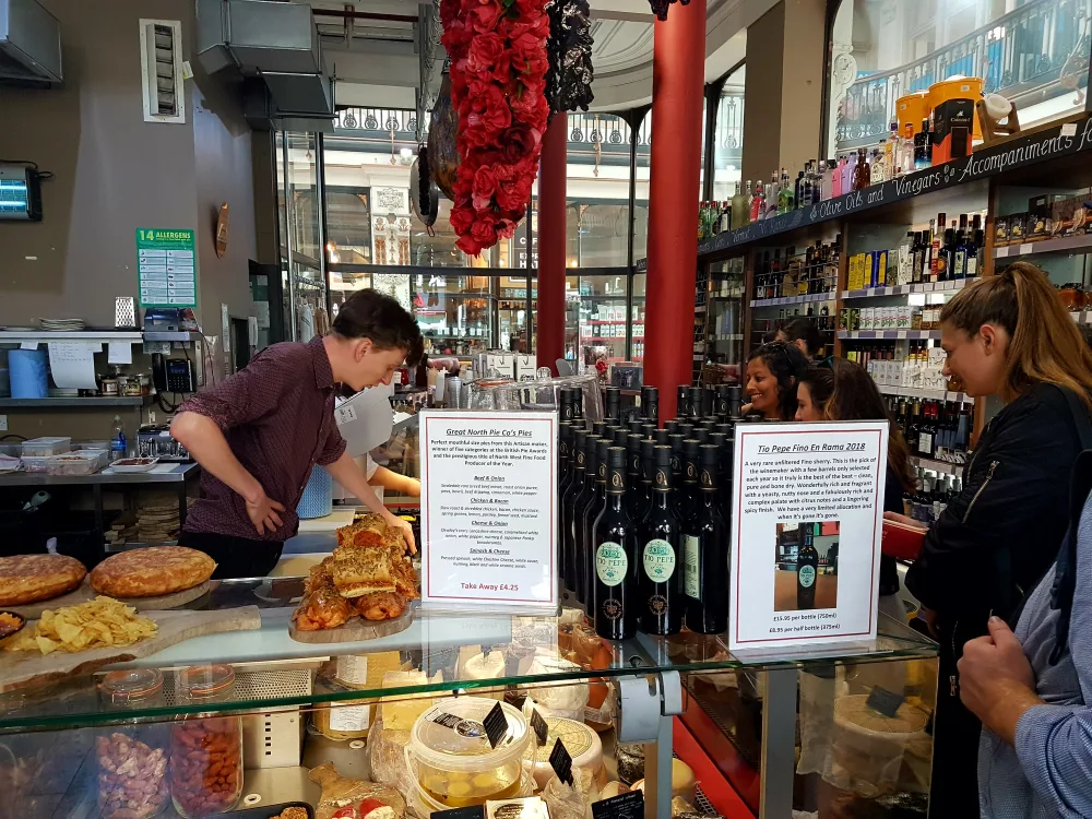 A man organizes sandwiches and confectionary from behind a till as customers peruse the offering, at a deli