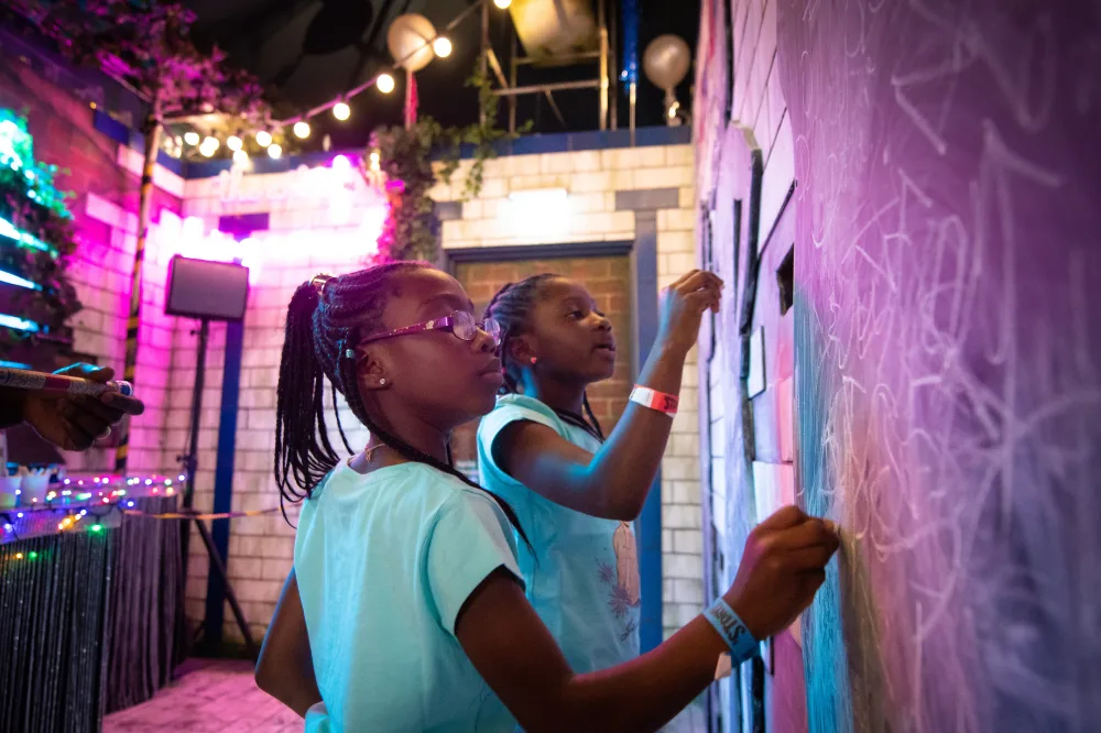 Two young girls draw on the chalk board wall with colourful chalk. Twinkly lights sit in the background.