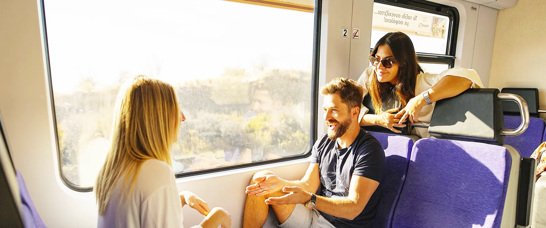 Two women and a man chat onboard a train in Europe, from the window you can see blue skies and scenery on a hot day