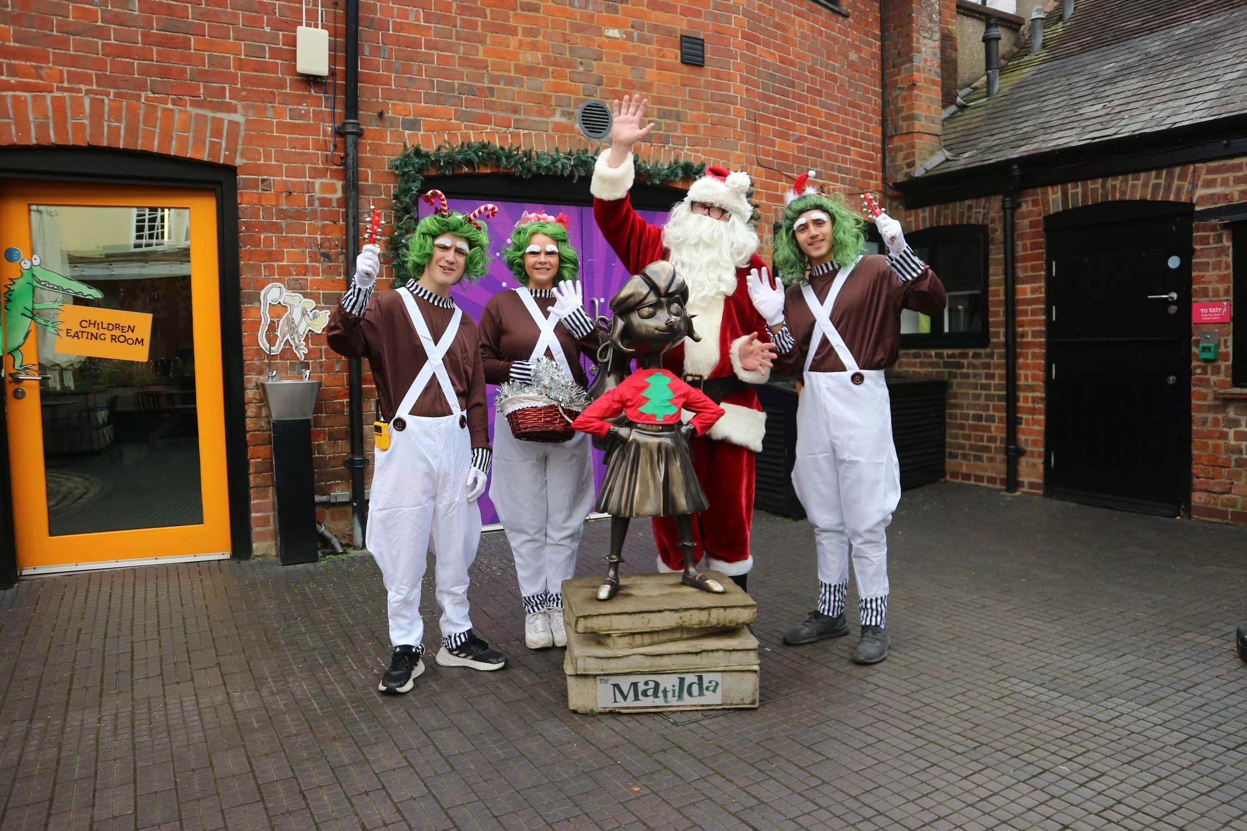 Performers in Santa and Oompa Loompa costumes posing with a Matilda statue outside a brick building.