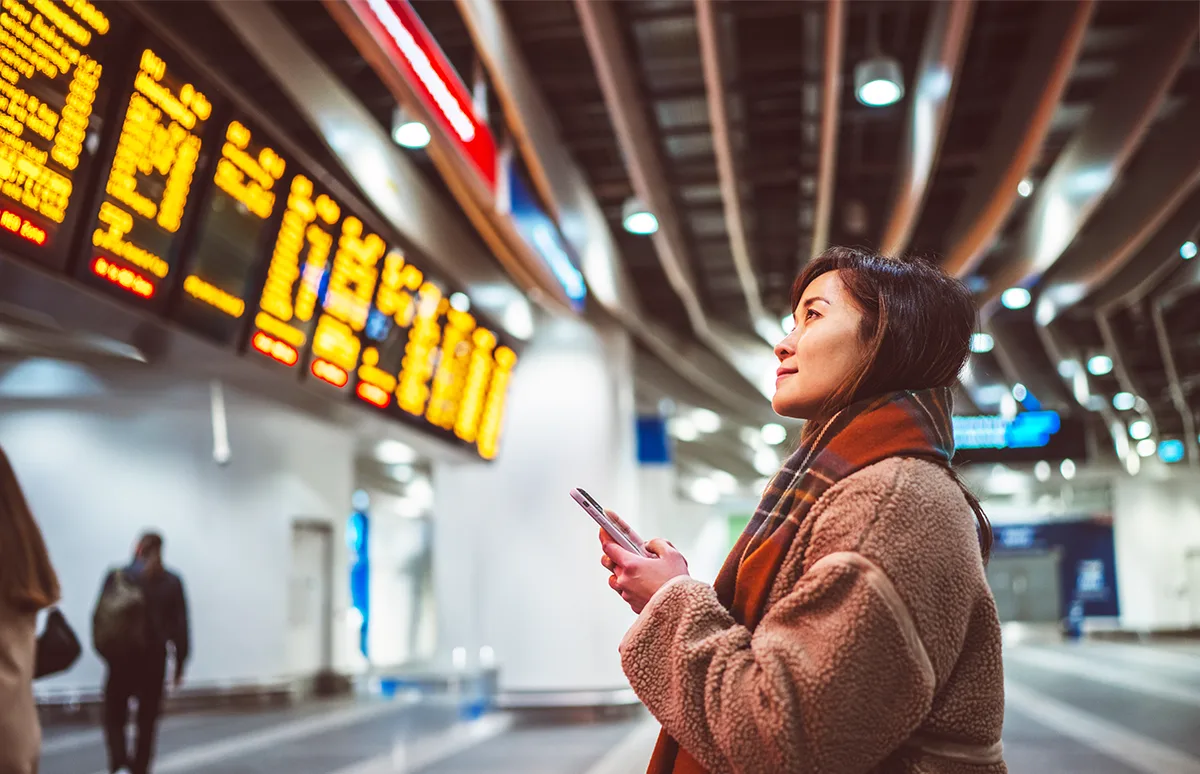 A woman in a tan coat holding a smartphone looks up at illuminated departure boards in a busy train station.