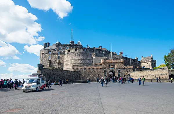 Edinburgh castle, a large grey stone building, on a sunny day, with scattered groups of people walking towards it across a paved area. 
