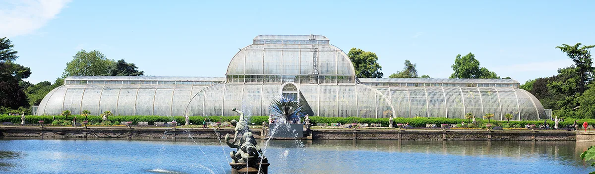 A large greenhouse next to a lake, with a statue and fountain in the middle of the water. 