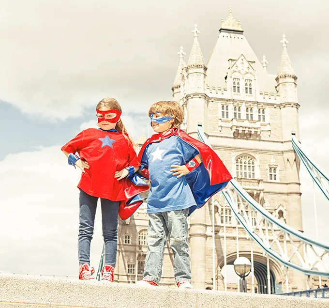 A white girl and boy in superhero masks and capes stand in front of Tower Bridge.