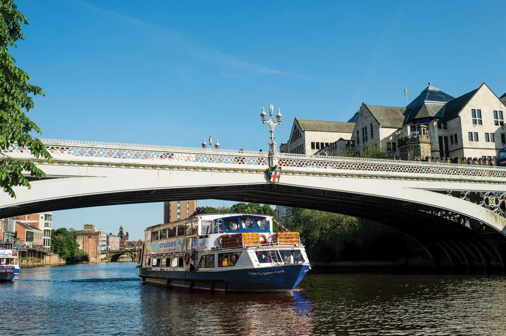 A river cruise boat going underneath a white stone bridge on a sunny day.