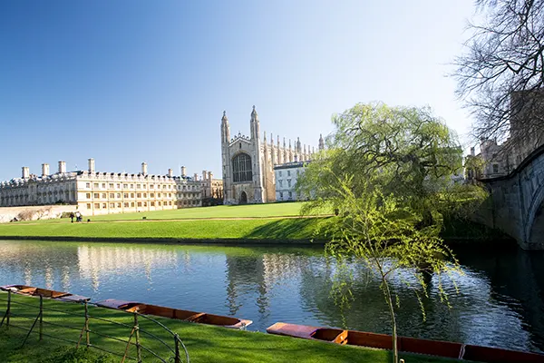 An ancient cathedral and university buildings on a manicured green lawn leading down to a river with wooden punts moored at the edge. 