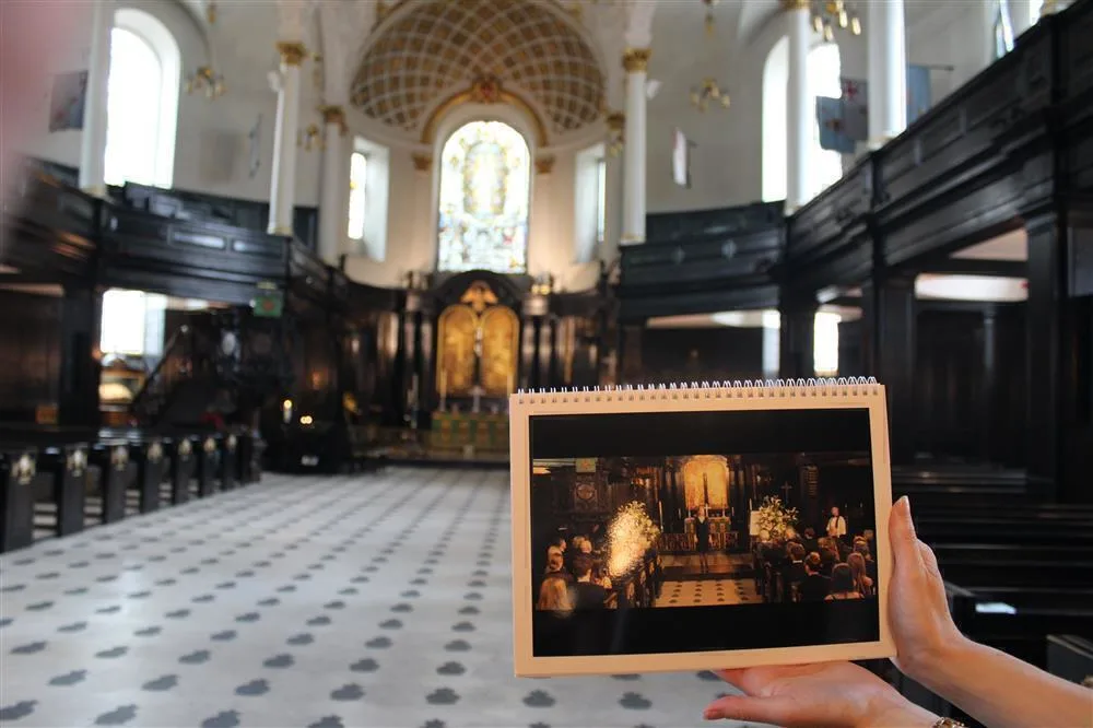 Hand holding a photo of a wedding ceremony up against the actual background in the historic church with white walls, dark wood panelling and stained glass.