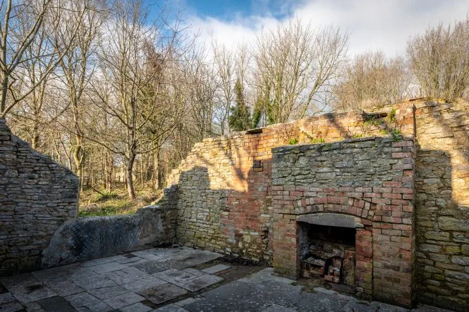 Old ruined building comprised of brick with an old open fire place sits beneath a cloudy and blue sky with trees overlooking the site.