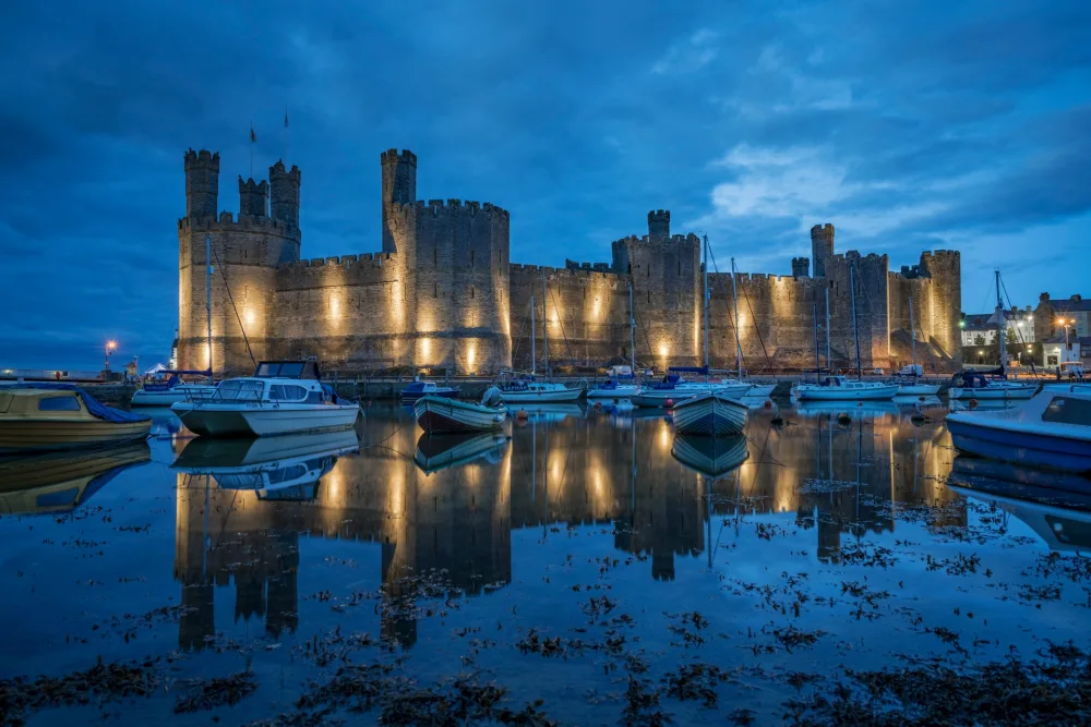 An historic Welsh castle illuminated at night and reflected in the river in front. 