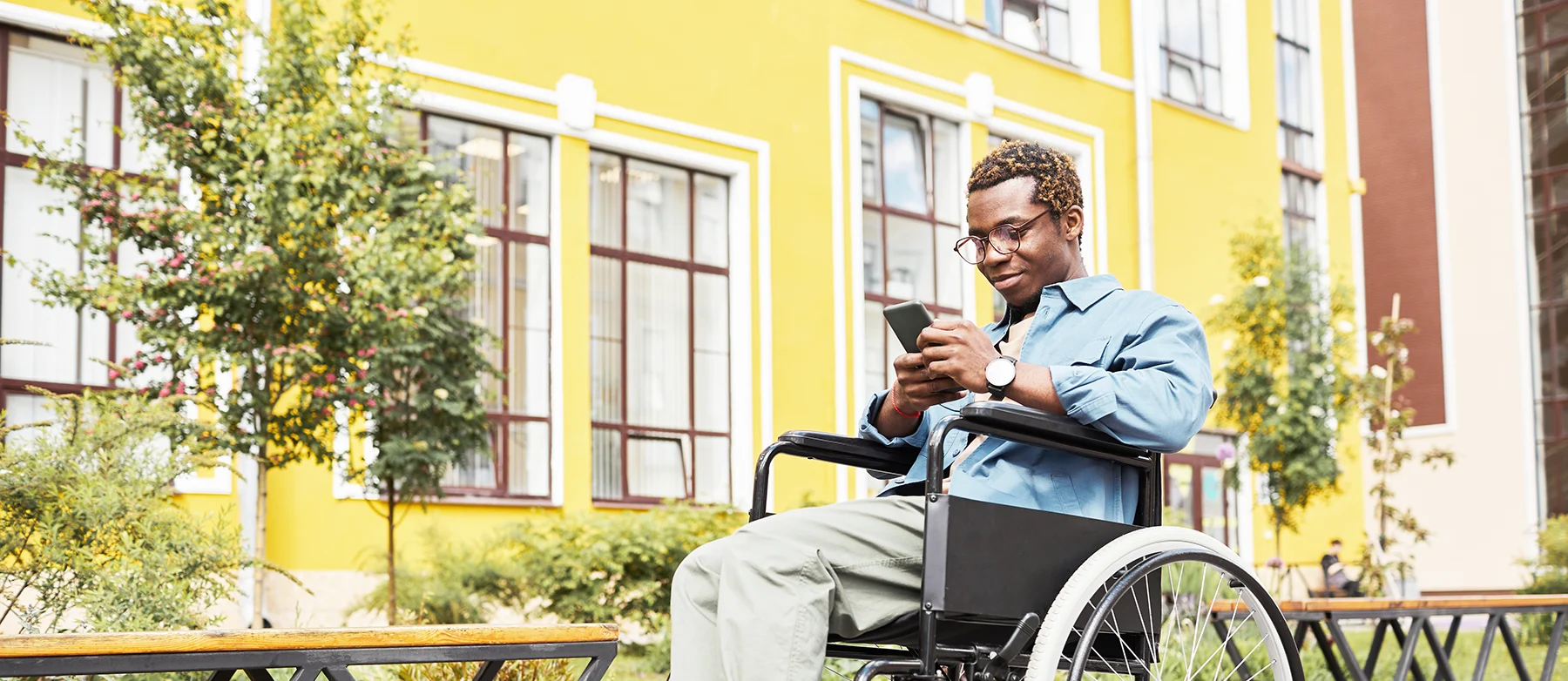 A young casually dressed Black man in a wheelchair looking at a mobile phone, on a pretty tree-lined street 