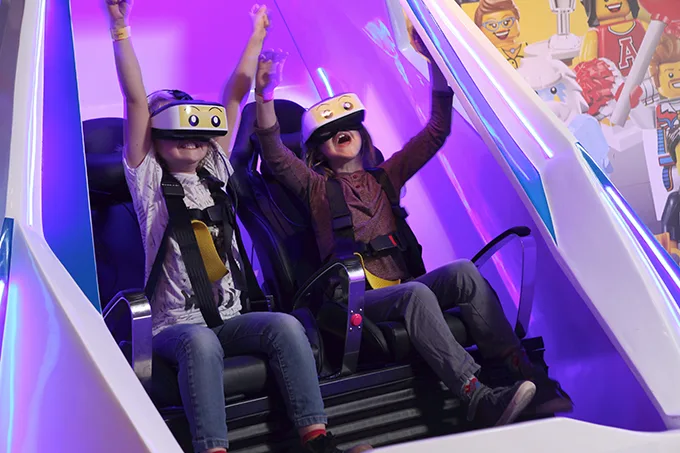 2 children sitting on a ride, wearing virtual reality goggles and lifting their arms in the air.