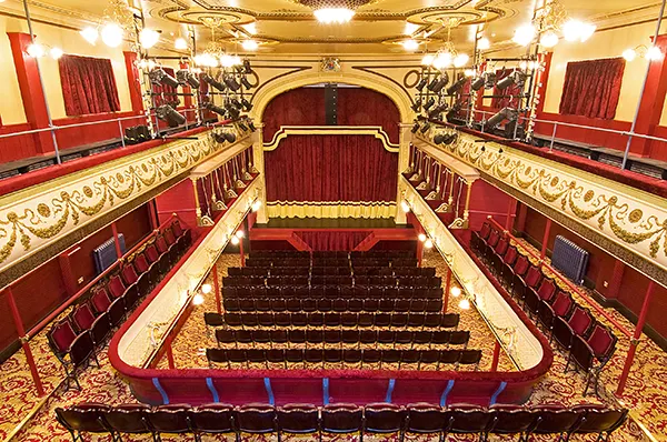 View from the circle of an empty variety hall theatre, the stage has a large red curtain and the seats are red velvet.