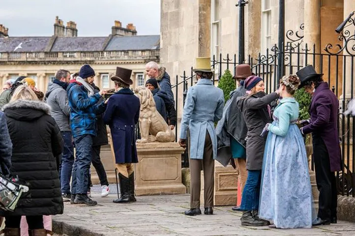 Actors dressed in period attire of top hats and full length gowns get touch ups from film crew outside a grand building. 