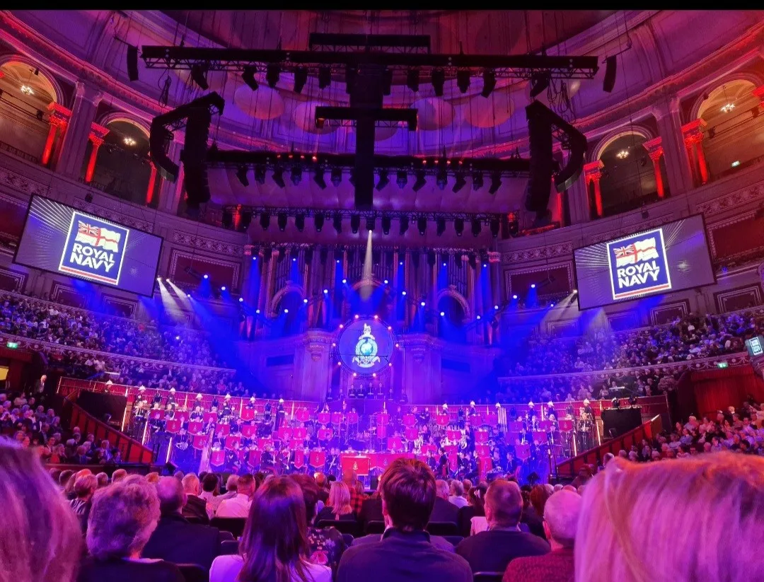 An audience view of a military band playing on stage under lights and screens. The words 'Royal Navy' appear on screens to the left and the right of the stage.
