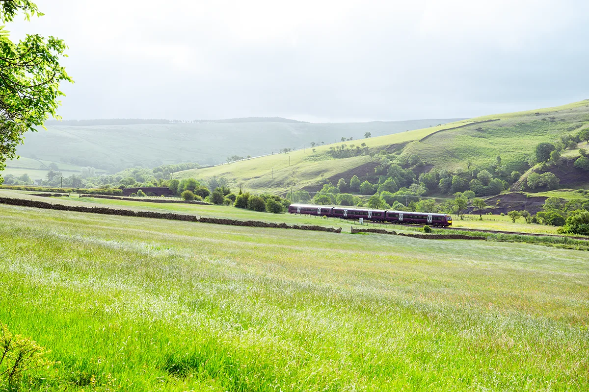 A passenger train travelling through lush green rolling hills of the British countryside under a misty sky.