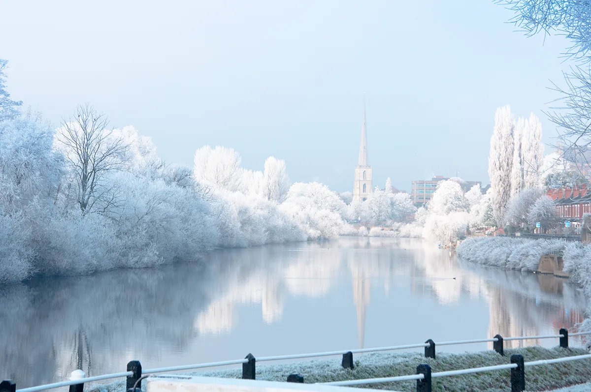 A still river surrounded by snow-covered trees with a cathedral visible in the distance.