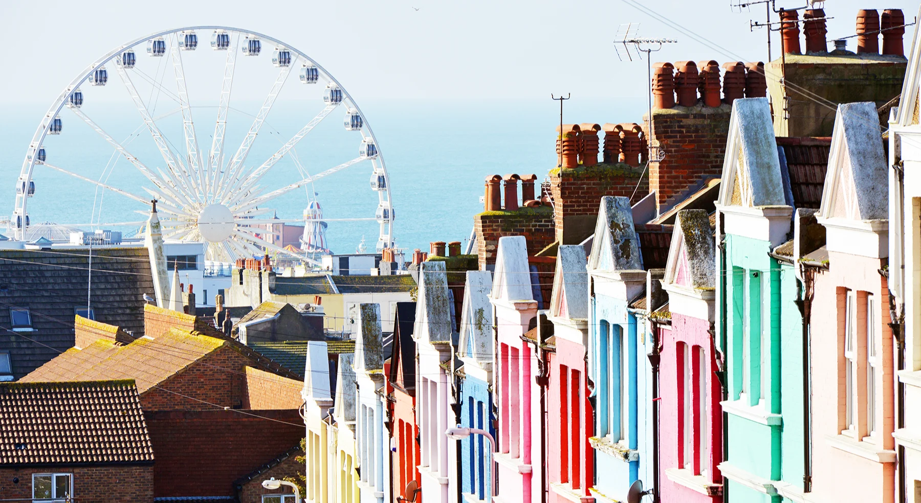 A row of brightly painted terraced houses with a view of the sea and a ferris wheel in the background.