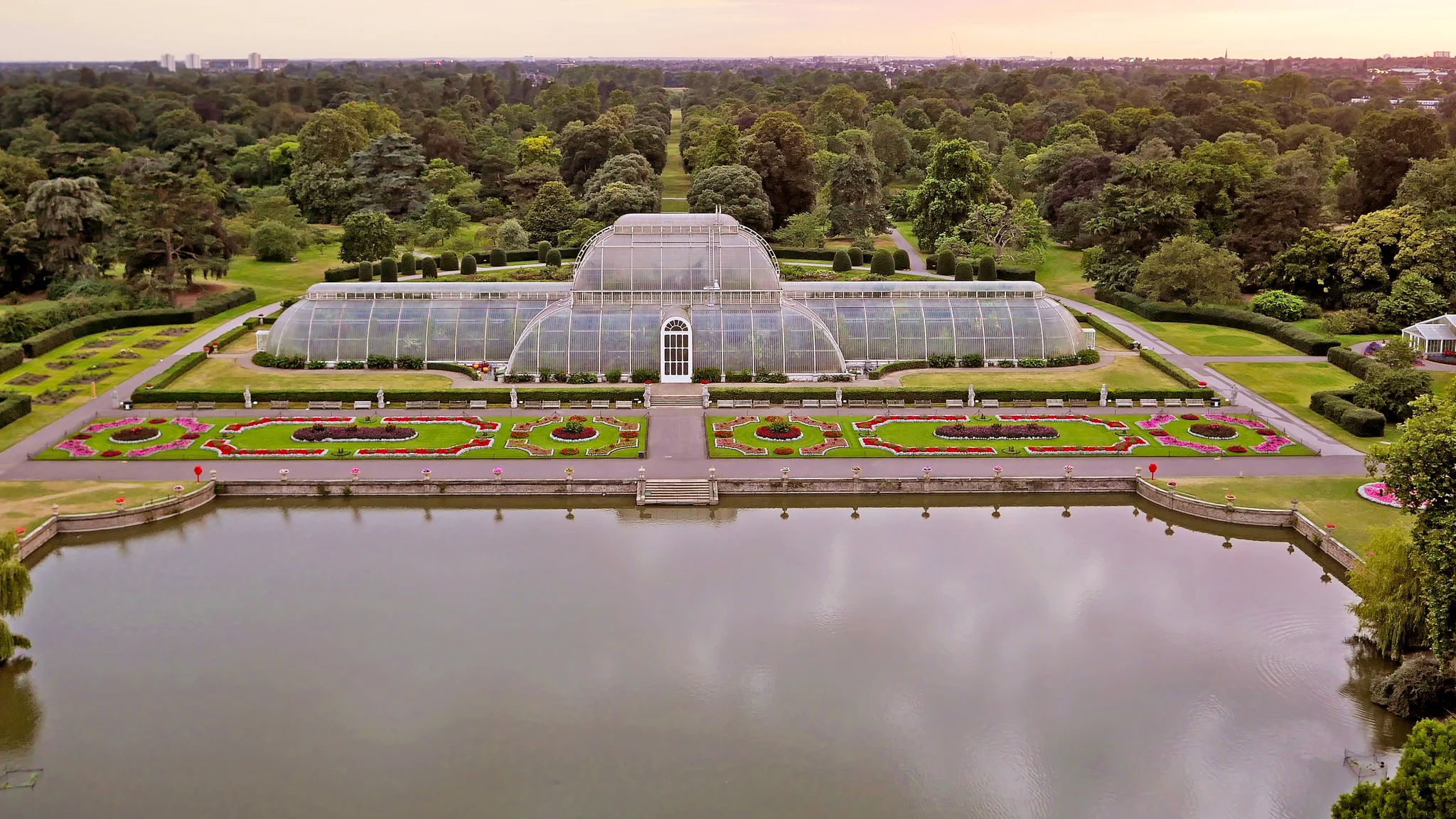 Aerial view of Palm House at Kew Gardens. Large Glass House sits within a well manicured garden, in front of large lake