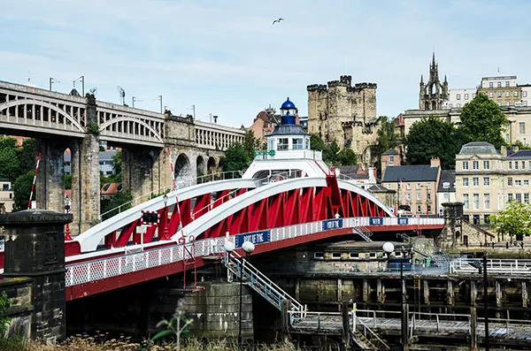 A red and white bridge over a river with a view of Newcastle's historic buildings in the background. 