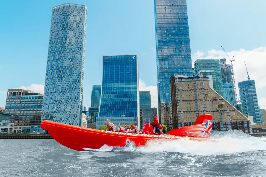 A red speedboat with passengers holding their arms in the air races down the river Thames in London on a sunny day with glass skyscrapers in the background.
