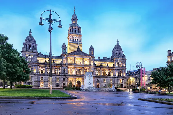 An imposing municipal building with tall spires and domes, lit at twilight, overlooking a pedestrian square. 