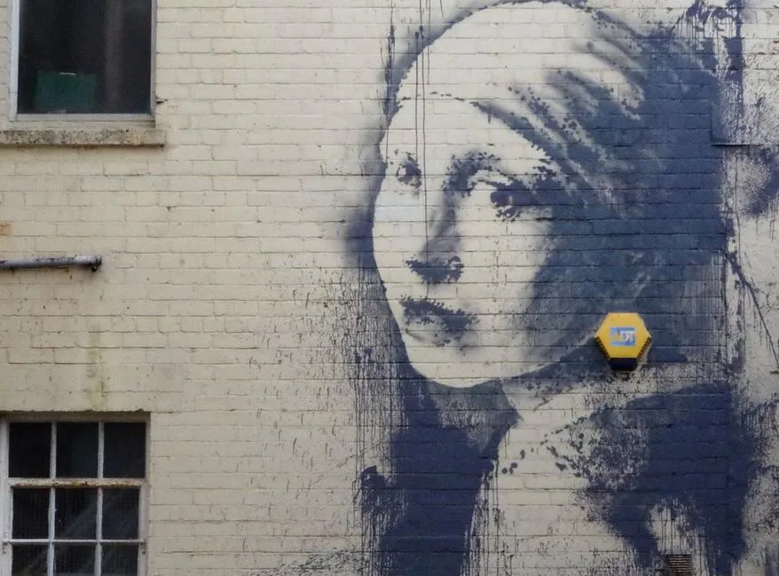 Street art by Banksy, based on the portrait The Girl With The Pearl Earring, painted on a white brick wall.