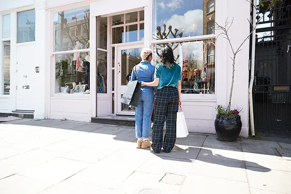 A Black woman with short bleached hair and a white woman with long dark hair, both are looking in a shop window and holding shopping bags. The white woman has her arm around the Black woman's waist. 