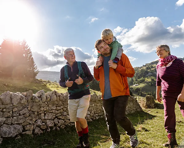 A family hiking outdoors on a sunny day, with a man carrying a young child on his shoulders beside a stone wall.