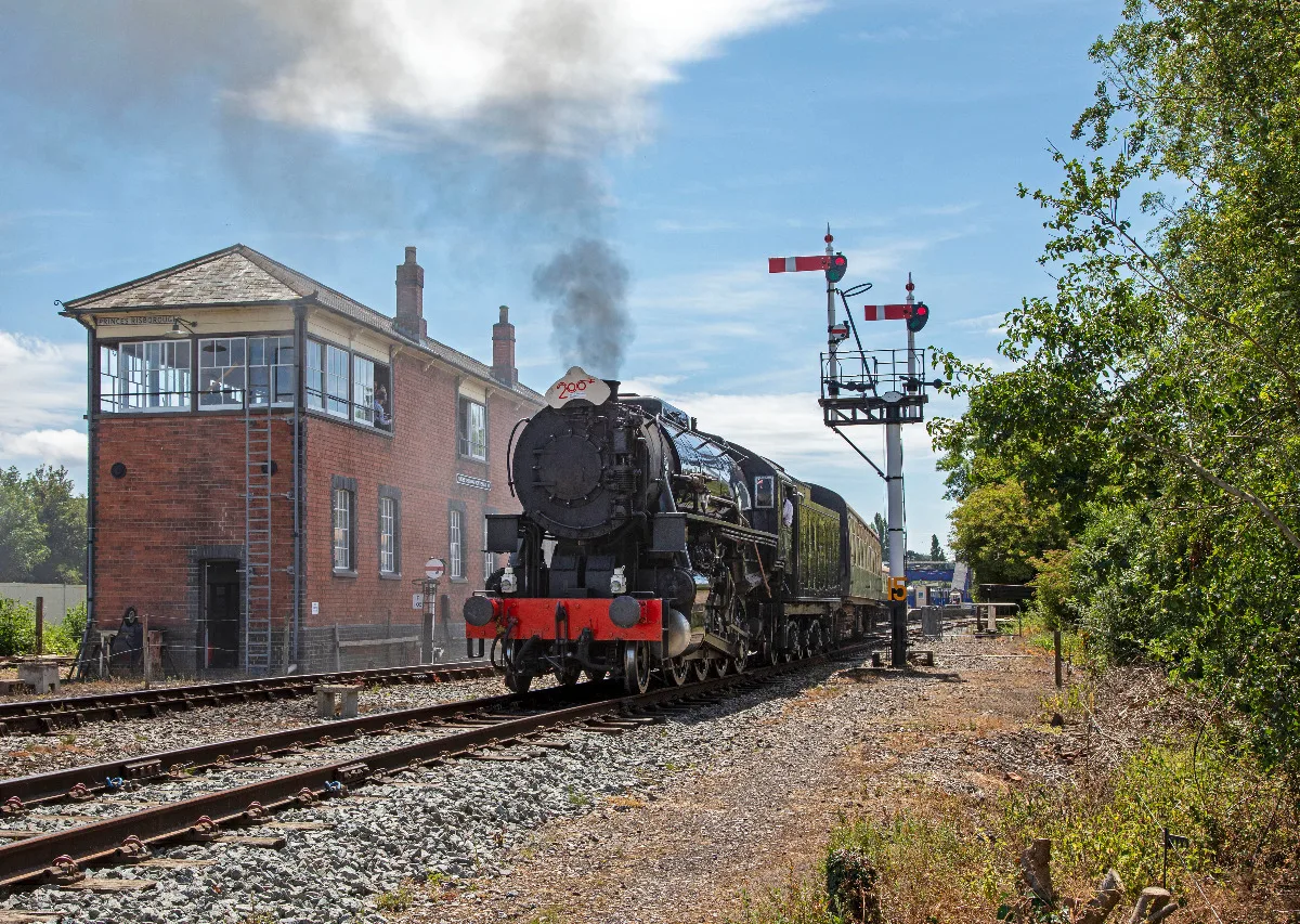 A green steam train coming towards the camera on a bright sunny day with a red brick building to the left of the tracks.