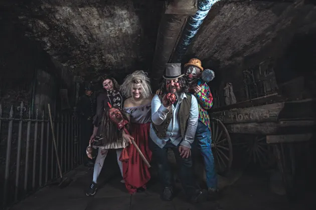 4 actors in spooky costumes looking at the camera in a sinister fashion, inside a dark underground tomb.