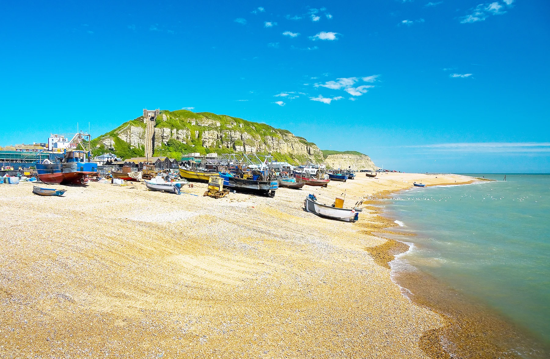 Looking along a shingle beach with fishing boats along the shore, a clifftop with a cliff railway is in the background, against a bright blue sky. 