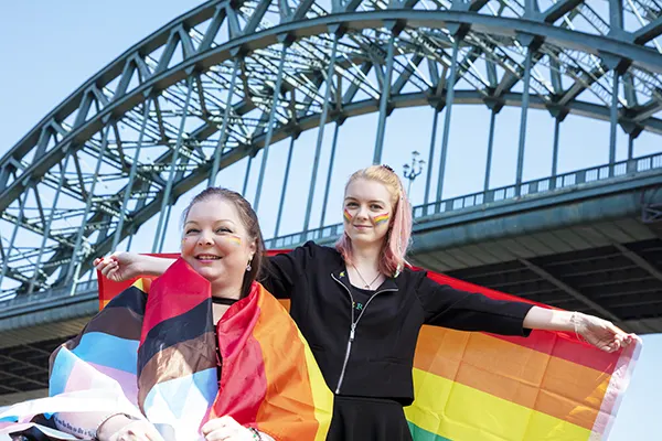 2 white women wrapped in gay and trans pride flags and with rainbow stripes painted on their cheeks.