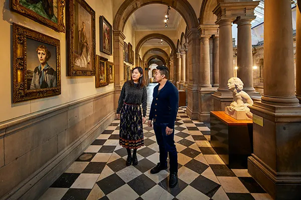 An East Asian man and woman standing on the black and white checked floor of an art gallery looking at a painting.