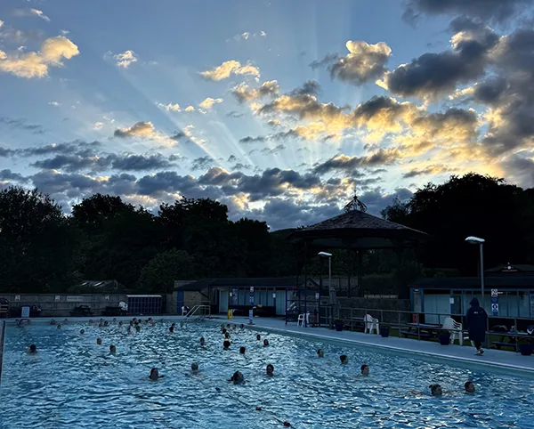 Swimmers in an outdoor lido at dusk, with dramatic golden sun rays breaking through dark clouds above silhouetted trees.